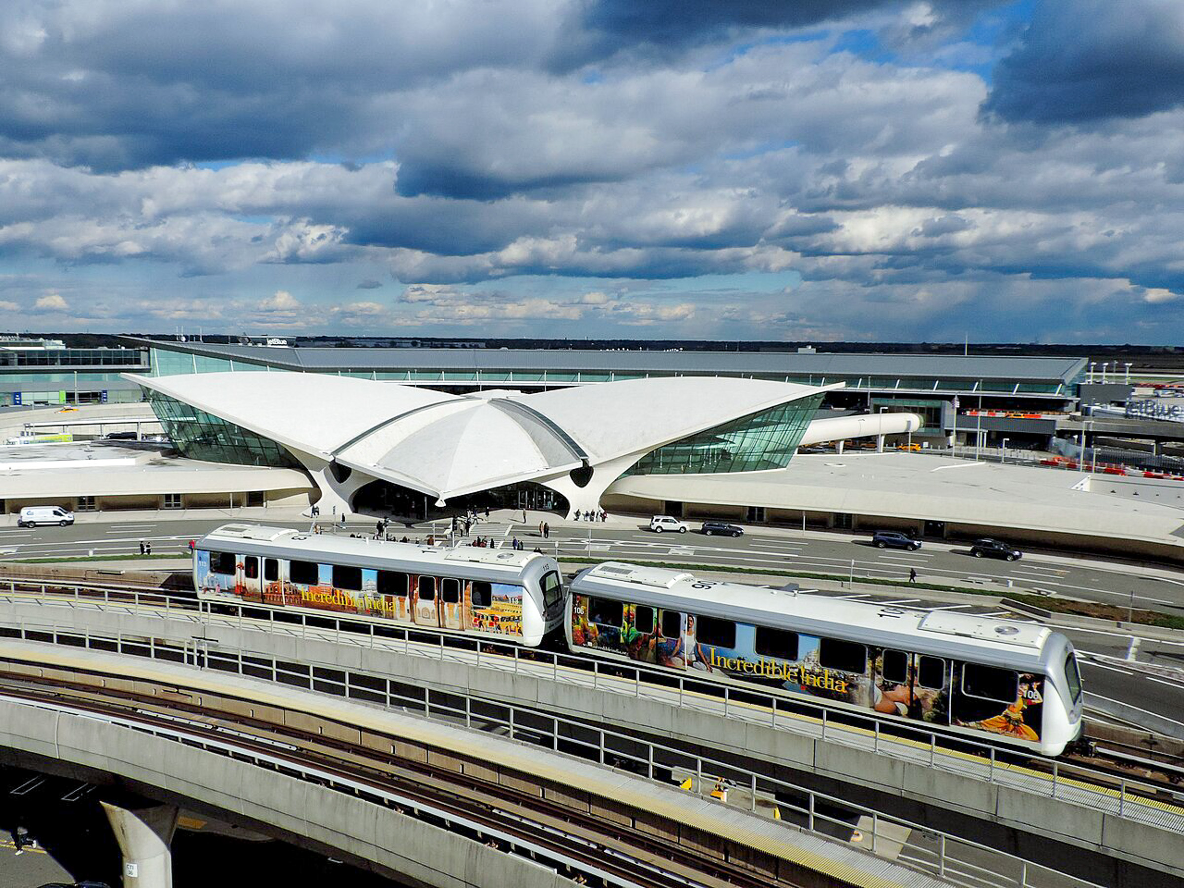ขบวนรถไฟ AirTrain JFK สีขาวคาดแถบสีน้ำเงินและเหลือง กำลังวิ่งอยู่บนรางยกระดับ โดยมีฉากหลังเป็นท้องฟ้าและป้ายบอกทางบนทางด่วน